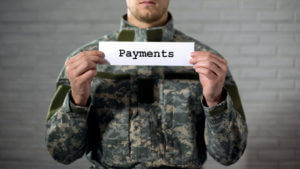 Payments text written on a sign held by a male soldier to illustrate veteran support, aid, and compensation.