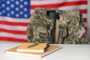 A notebook and pen are placed on a white wooden table, with a chair featuring a soldier's uniform against the flag of the United States, illustrating veteran compensation.